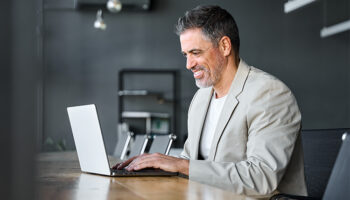 Homem de negócios de meia-idade ocupado e feliz, CEO, vestindo terno, sentado em uma mesa de escritório usando laptop. Homem de negócios maduro, profissional, executivo e gerente trabalhando em tecnologia corporativa no computador no local de trabalho.