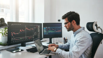 Jovem comerciante barbudo usando óculos, utilizando seu laptop enquanto está sentado em um escritório na frente de telas de computador com gráficos de negociação e dados financeiros.