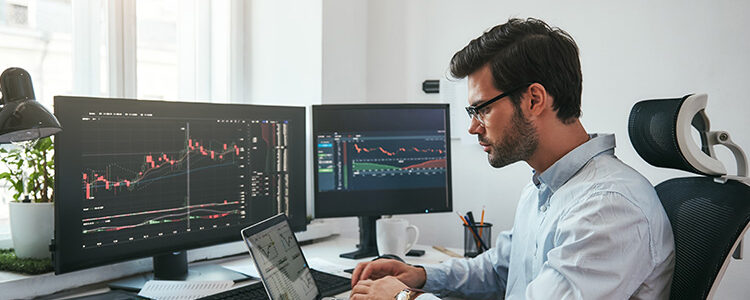 Jovem comerciante barbudo usando óculos, utilizando seu laptop enquanto está sentado em um escritório na frente de telas de computador com gráficos de negociação e dados financeiros.